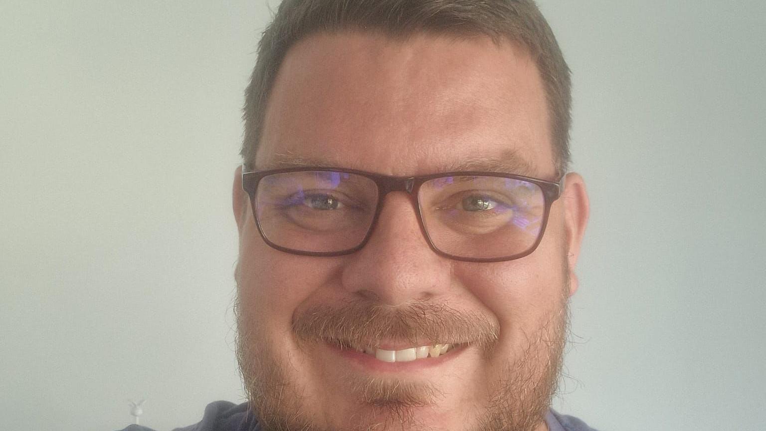 Close up of James, who is smiling and wearing glasses. He appears to be inside. He is unshaven and has short brown hair.