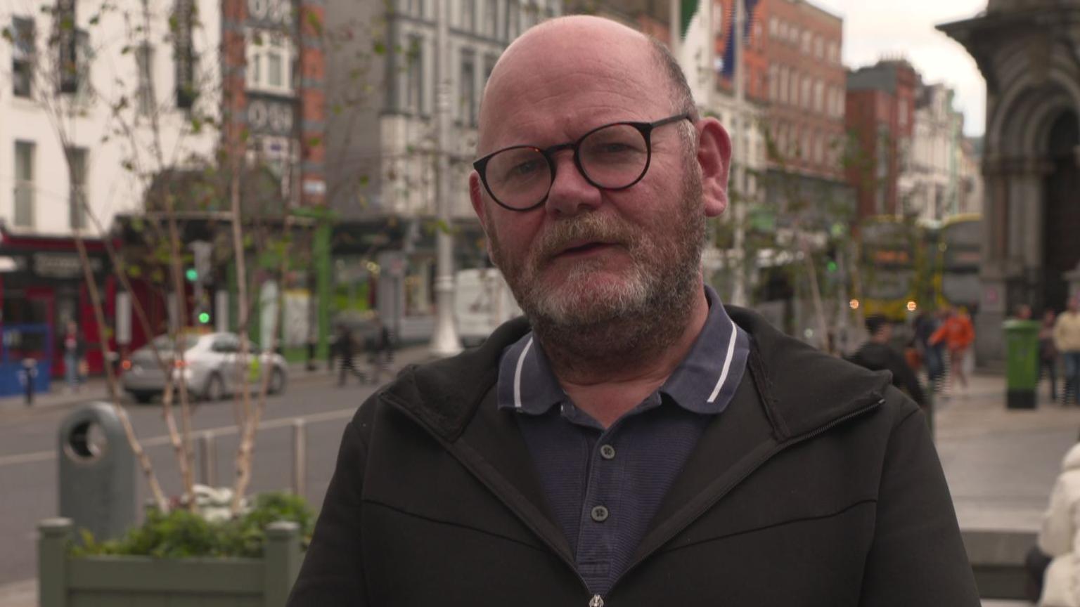 Daithi Doolan in the city centre of Dublin with cars and buses in the background, he is mostly bald with grey sides and a grey/brown beard. He is wearing black framed glasses, a blue/grey t-shirt and a khaki zip up jacket