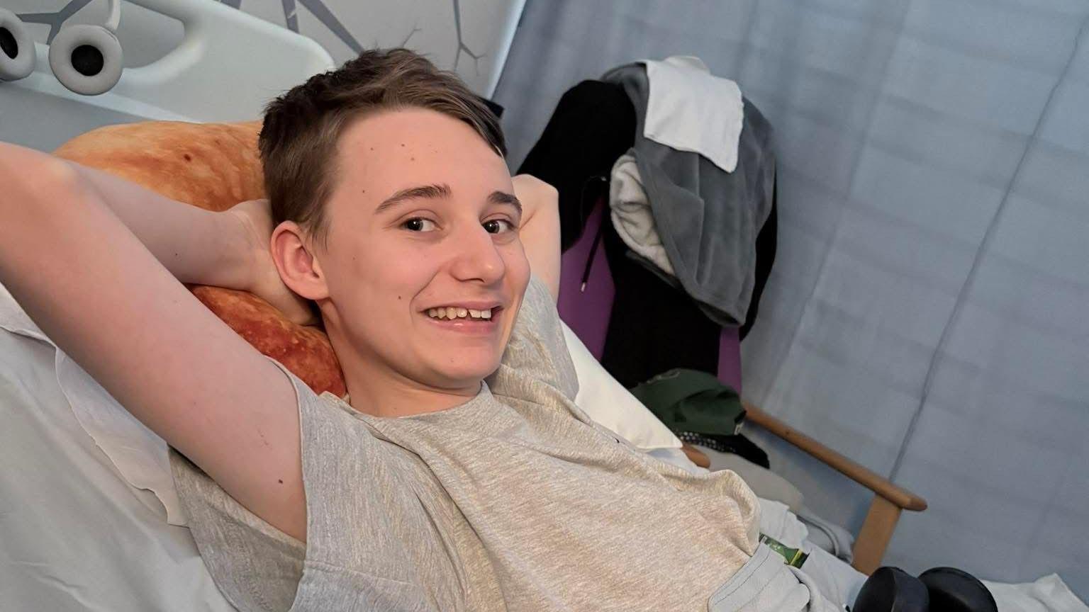 A teenage boy wears a grey short-sleeved t-shirt and relaxes in bed with his hands behind his head. He has a short brown hair. 