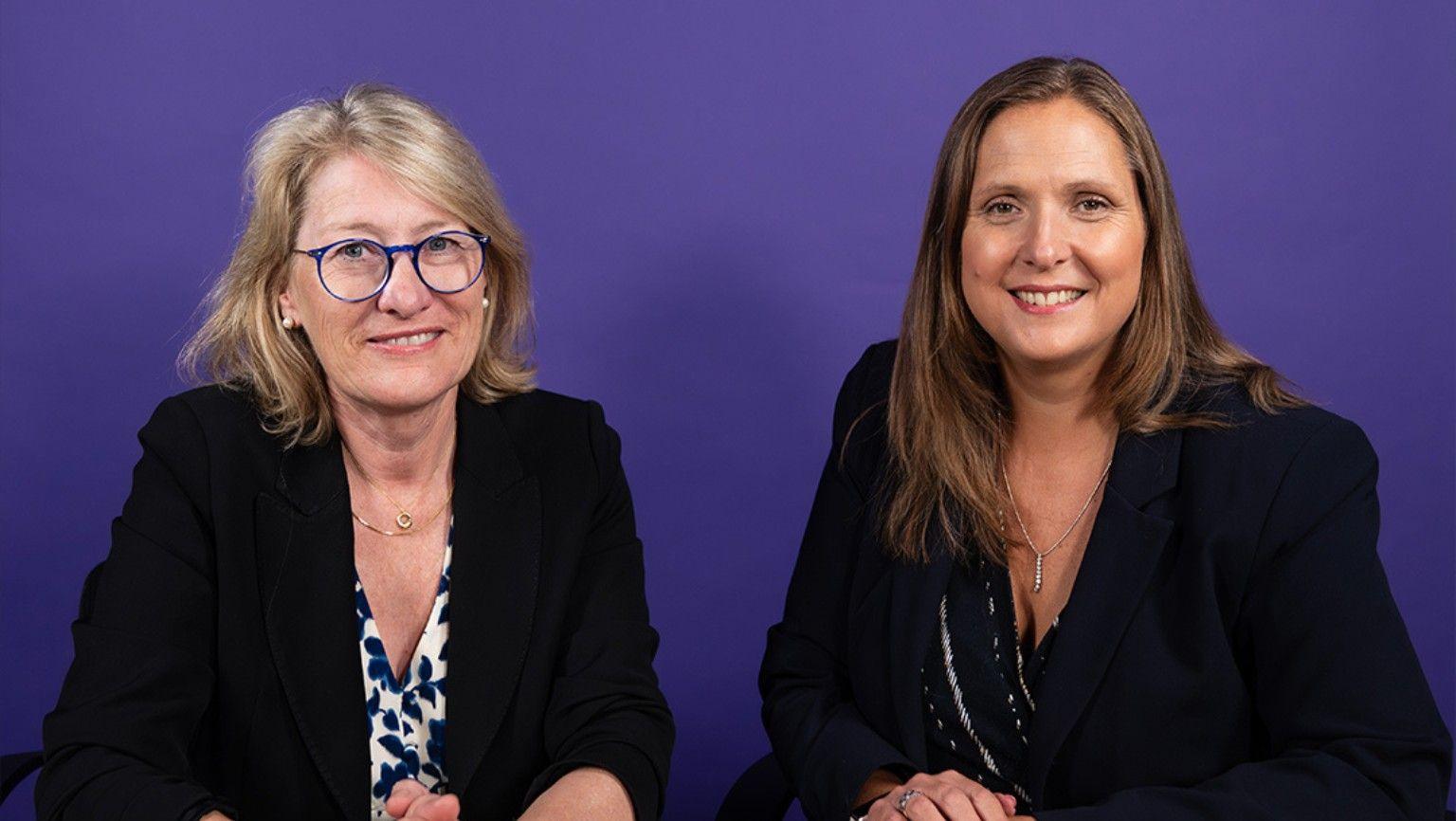 Two women sit side by side and smile at the camera. The woman on the left has blonde hair and is wearing a black blazer with a white and blue floral top. The woman on the right has brunette hair and is wearing a dark blazer and a dark top. The background behind them is purple.