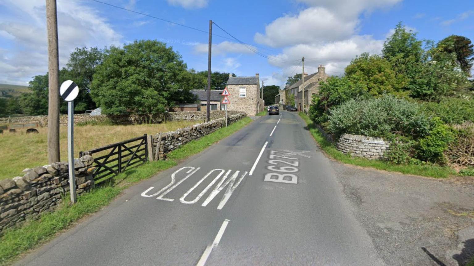 The B6277 in Newbiggin. It is a narrow two-lane road. To the left, there is a National Speed Limit sign and road markings reading SLOW. 