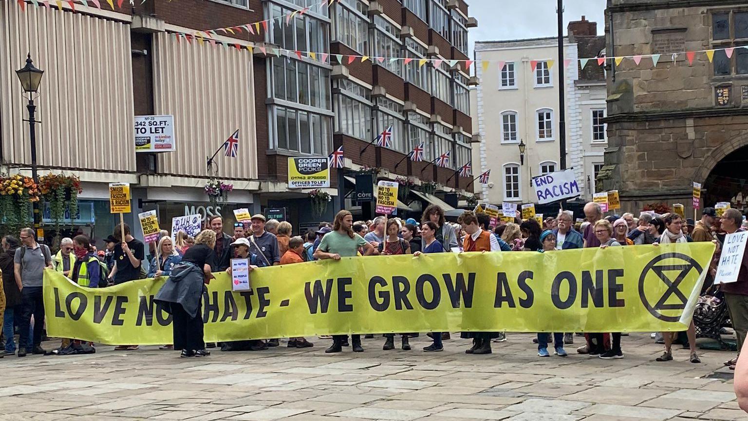 A group of anti-racism protesters with a large yellow banner reading "Love Not Hate - We Grow as One"