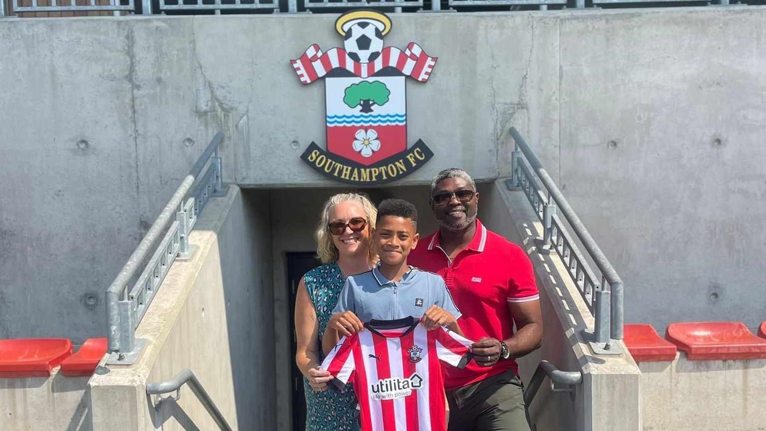 Kayo is stood holding a Southampton FC shirt with his parents behind him at a football stadium