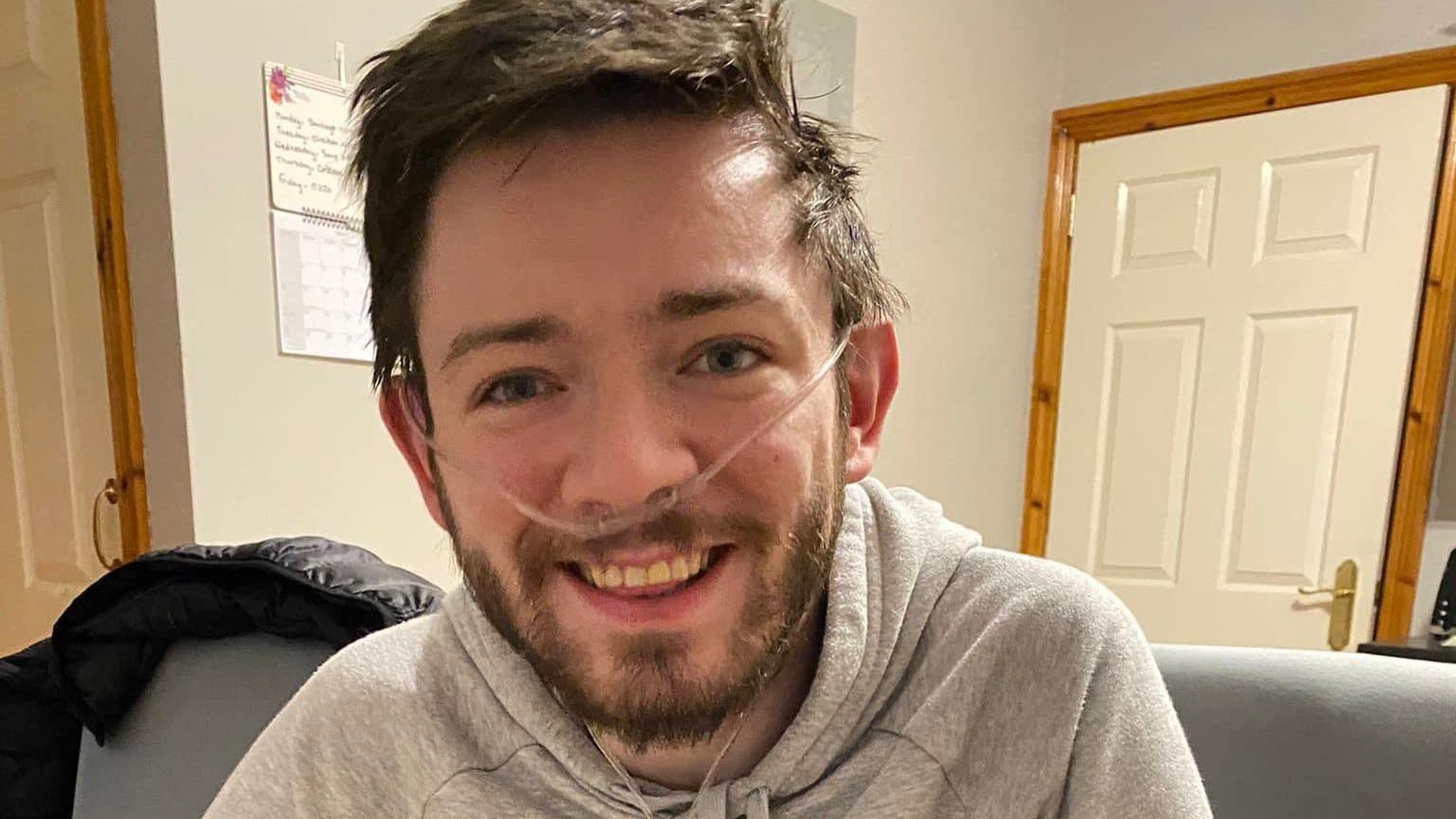 Man with short dark hair and facial hair, with medical tubes going around his ears and into his nostrils