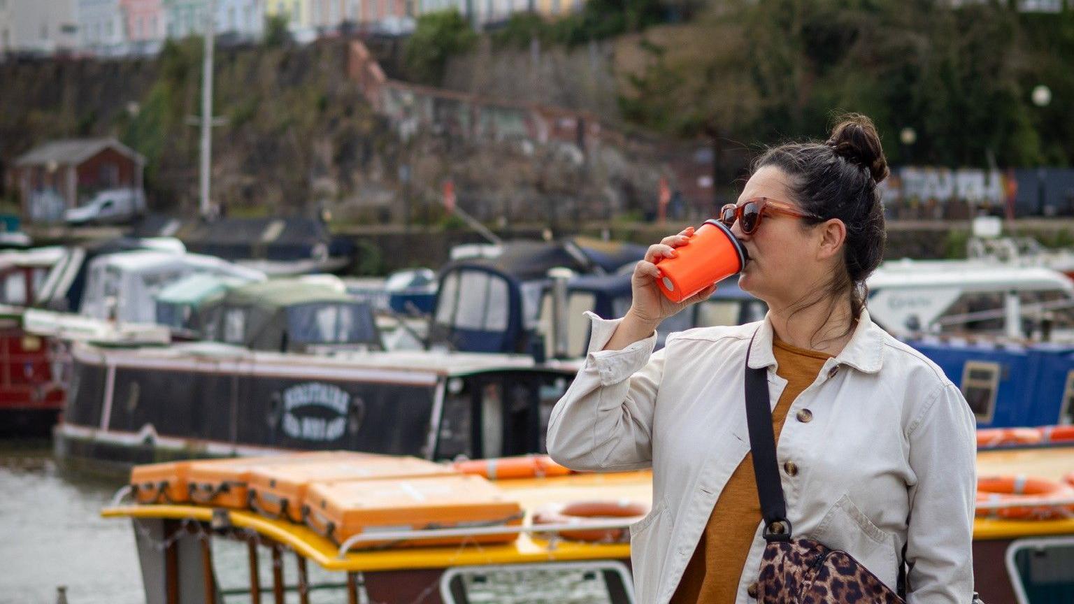 Elly looks off to the left of the frame as she drinks from a bright orange re-usable coffee cup. She has her brunette hair up in a bun and is wearing orange sunglasses. She has a white denim jacket on, an orange t-shirt and a small bag across her. Behind her canal boats are tied up in a marina. 