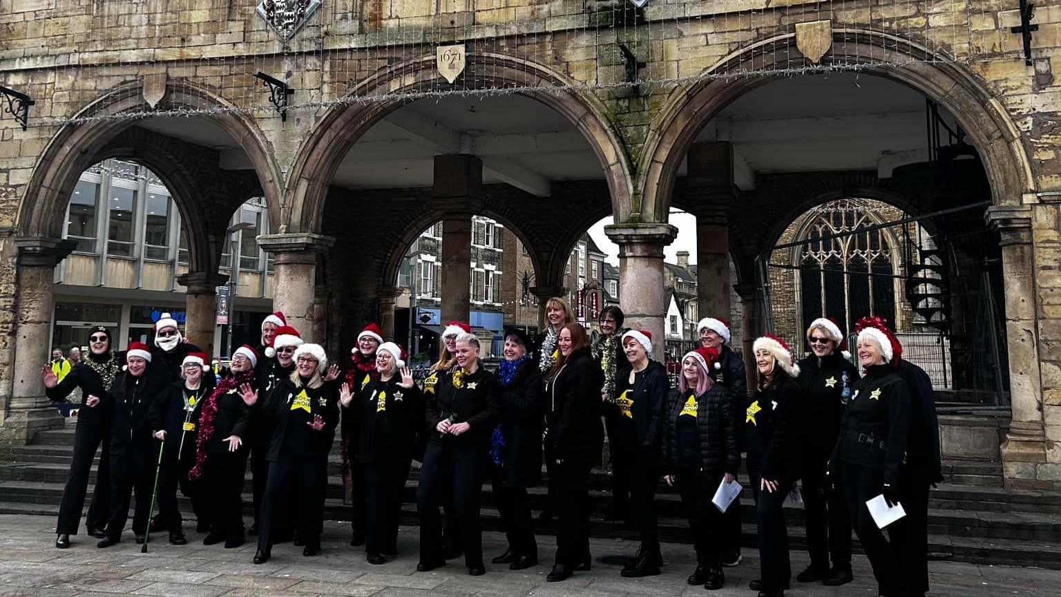 A choir dressed in black and wearing red Santa hats and white ribbons singing in front of the arches of a guildhall 