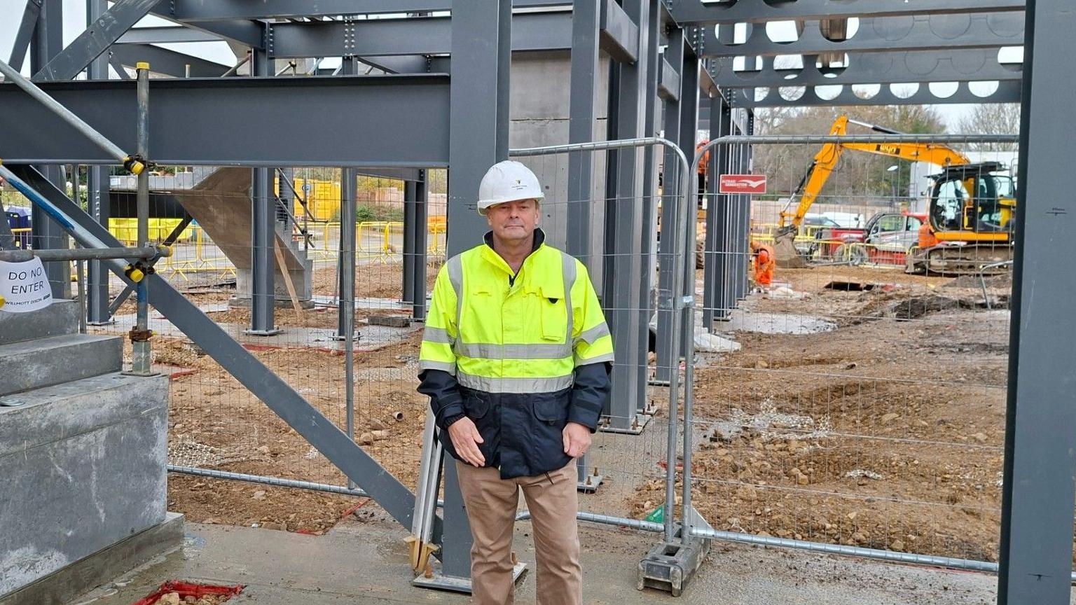 Man in white hard hat and high-visibility jacket stands on muddy ground at a construction site, facing a steel framework structure with beams and columns; excavator and building materials are visible, with a "Do Not Enter" sign on a staircase and temporary fencing surrounding the area.
