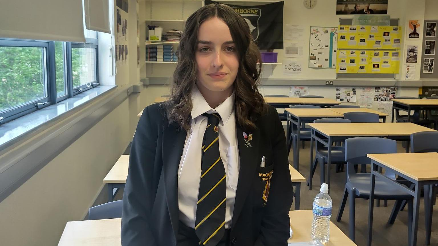 Katy is standing in a classroom looking at the camera. She is wearing a school uniform