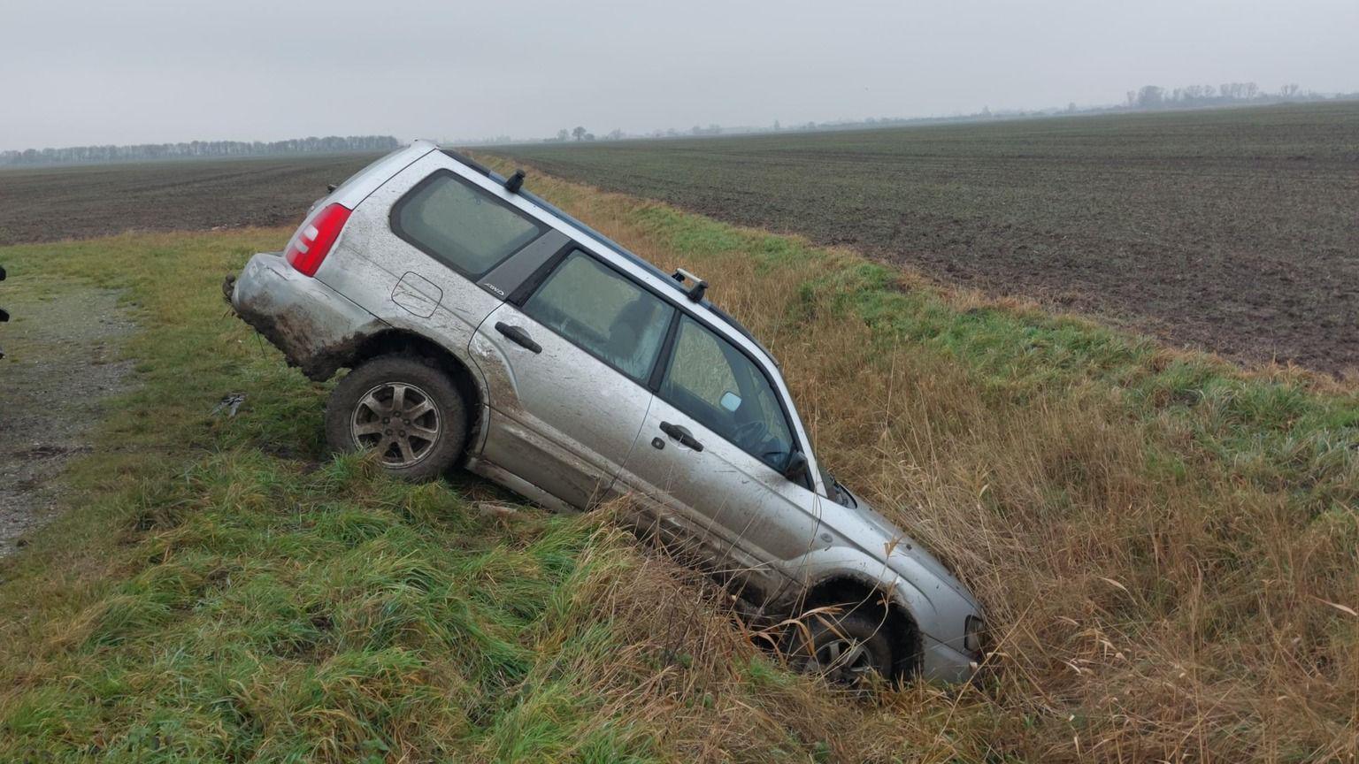 A silver car has nosedived into a ditch. The back wheels are at the top of a mound of grass and the front of the car is touching the ground. The surrounding area looks to be fields used for farming. 