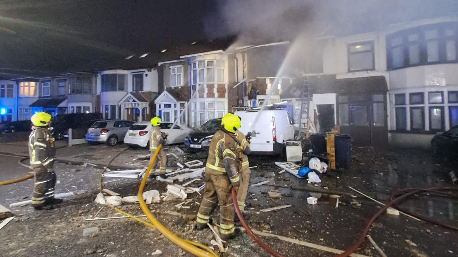 Firefighters in yellow helmets are working at the scene of a house fire at night, with smoke billowing from a damaged terraced house. Debris is scattered across the street, and fire hoses are in use.