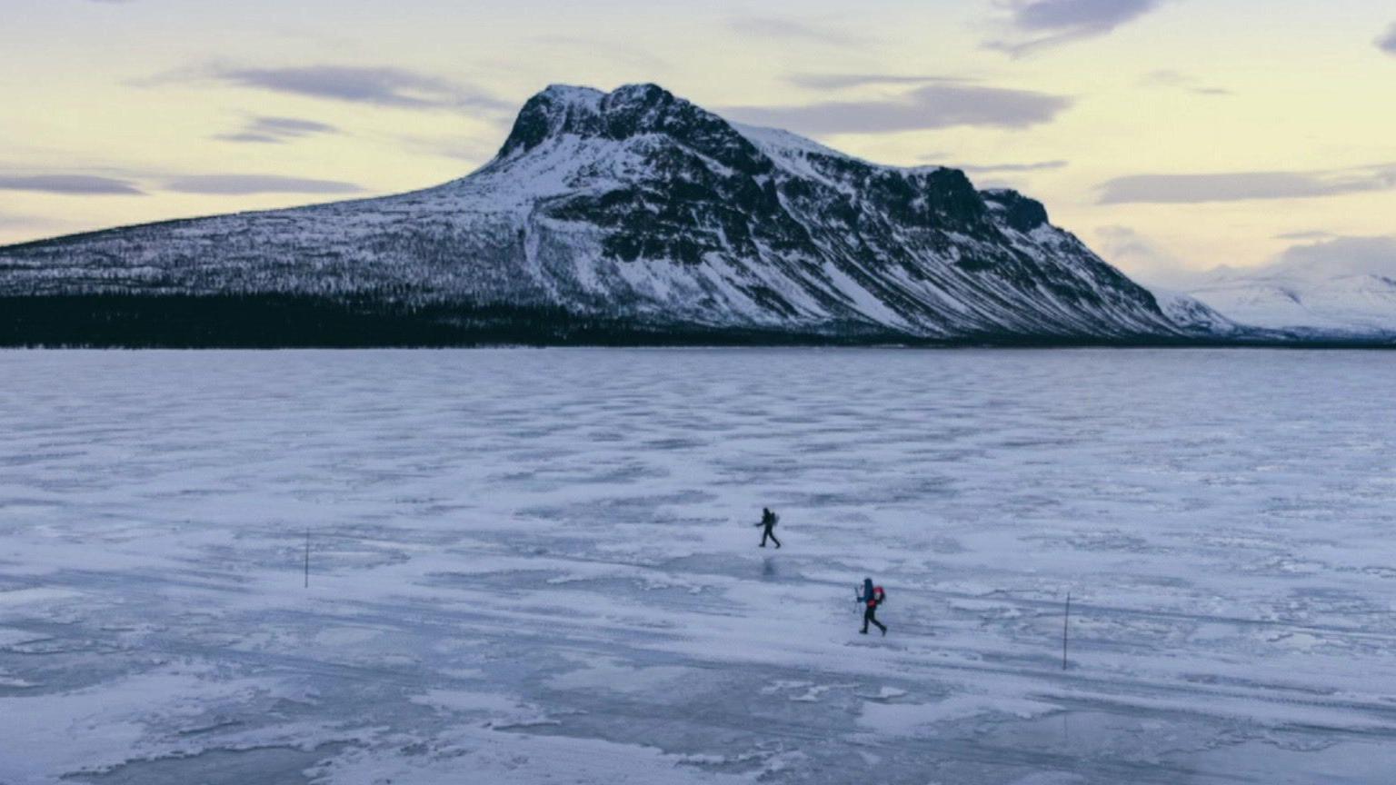 Two people run across the ice, they look tiny against the mountain in the background