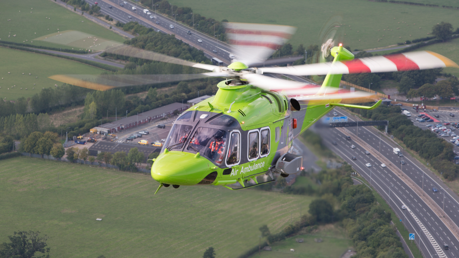 A neon green helicopter, with the words 'Air Ambulance' written on its side, flying over fields and roads. 