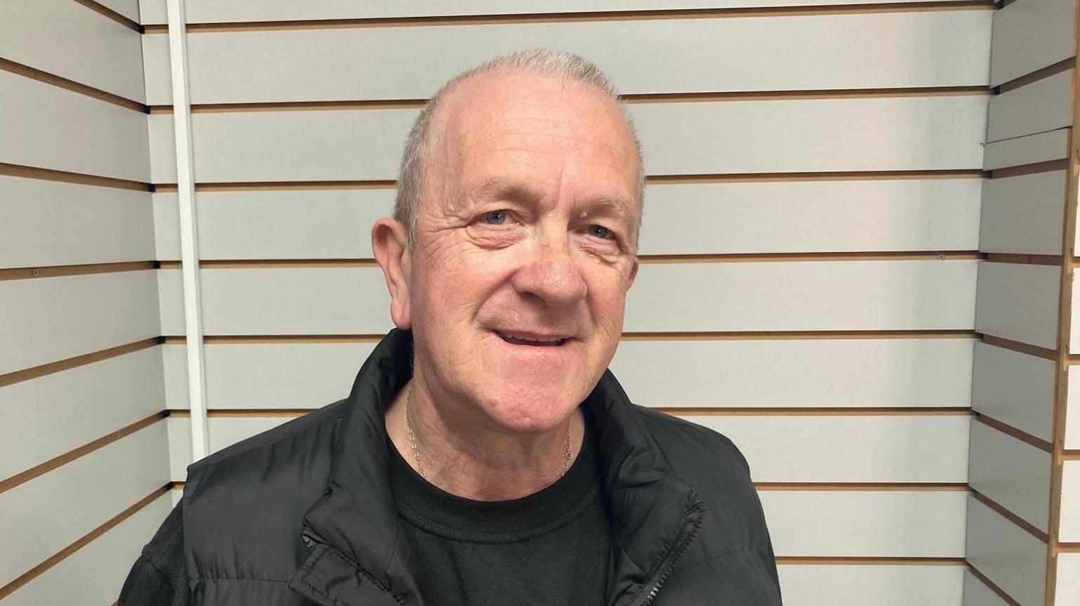 A man with thinning white hair looks at the camera with a slight open-mouthed smile. He wears a black jacket over a dark top and is in front of walls with thick white stripes broken up by thinner brown ones.