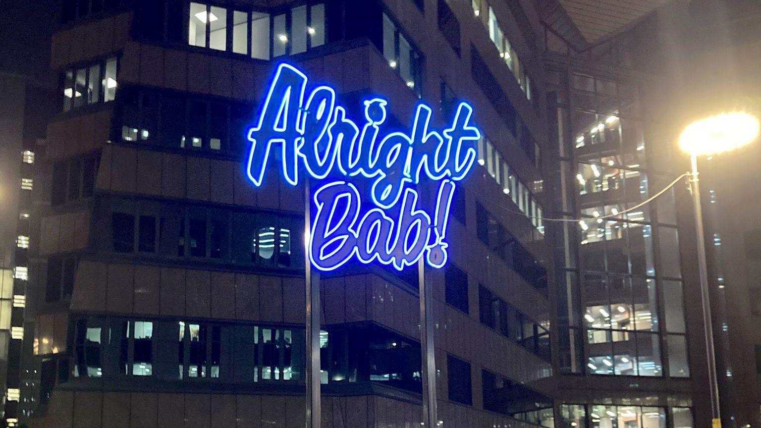 An illuminated blue sign in front of an office block reading Alright Bab, exclamation mark