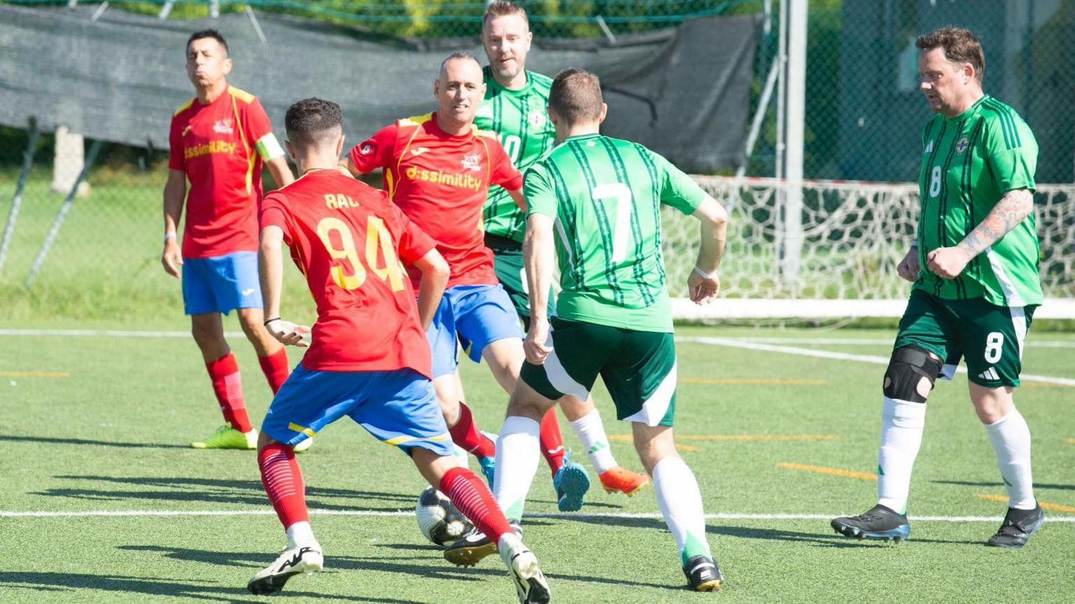 Six male football players on a football pitch with a football. Three of the men on the left are wearing red shirts with blue shorts. To the right, three men are wearing green shirts and shorts. 