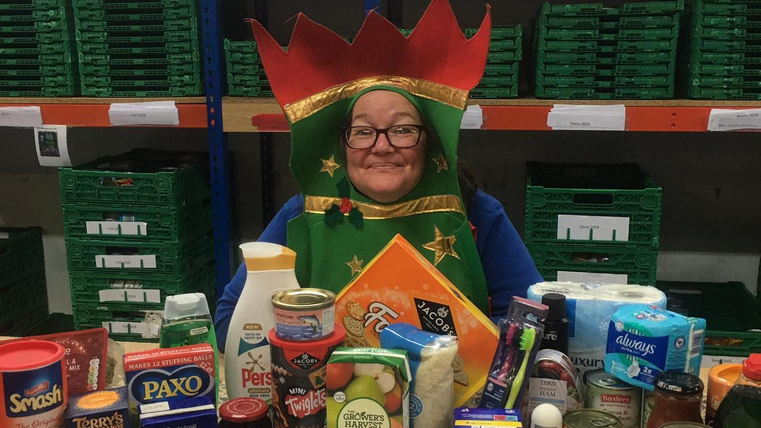 Maria Stevenson, a middle-aged woman in glasses, smiles at the camera while dressed in a Christmas cracker costume that surrounds her face. In front of her on a table is a selection of items: festive biscuits, stuffing mix and other food as well as essentials including toothbrushes and loo roll. There are stacks of green plastic crates behind her.