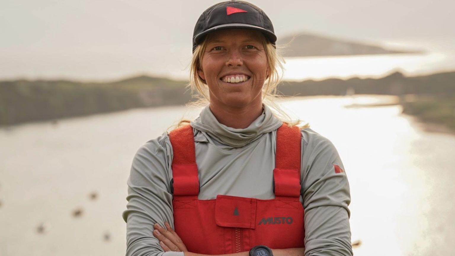 A woman with blonde hair and a black baseball hat stands looking straight at the camera. She is smiling with her arms folded. She is wearing a long sleeved grey jacket and a red Musto overall.