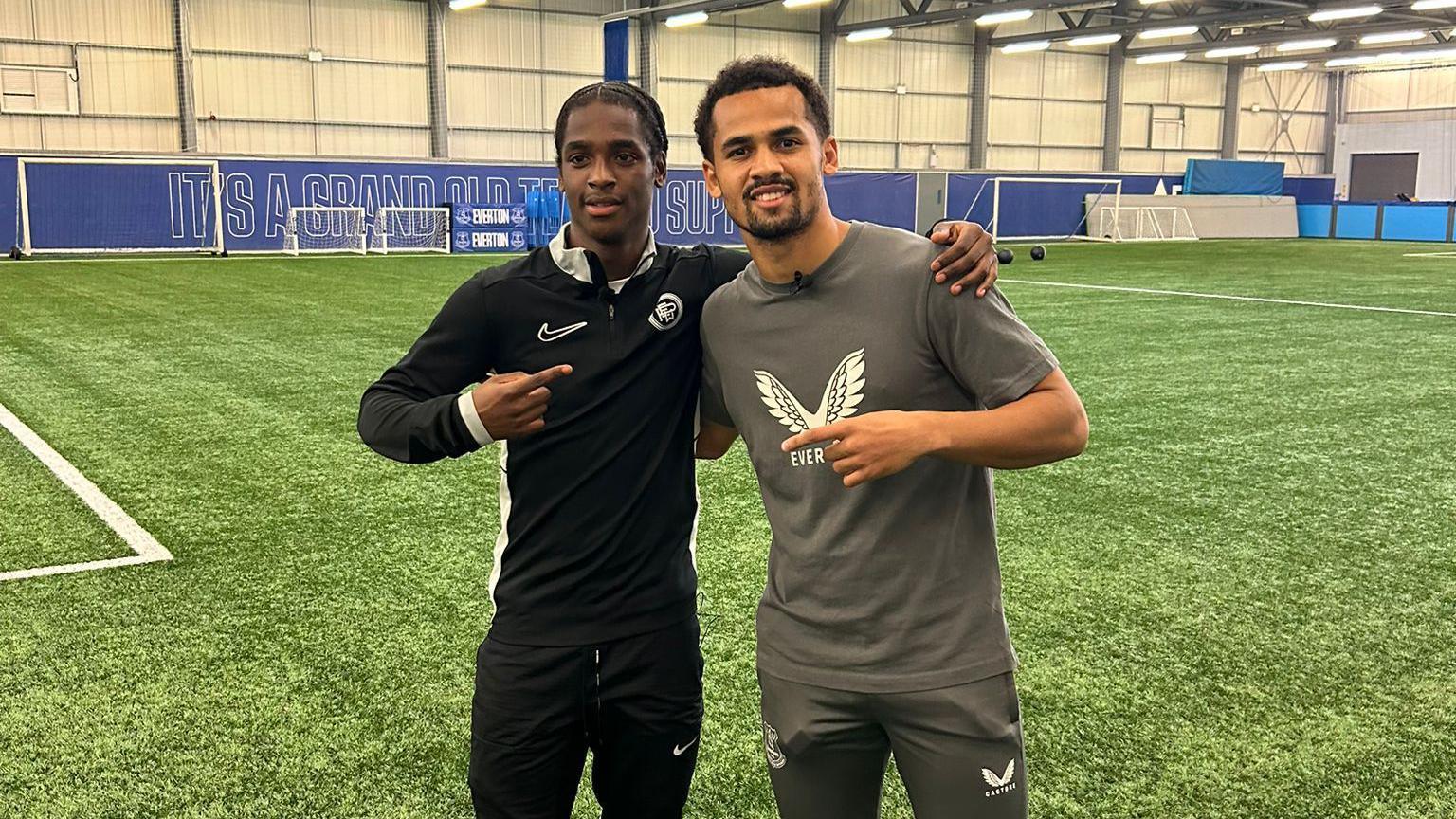 Kevar March-Mckenzie wears a black Nike tracksuit and he pointing to Iliman Ndiaye, who is stood beside him on a indoor football pitch. Ndiaye is wearing a grey t-shirt and tracksuit bottoms and is pointing back at Kevar.