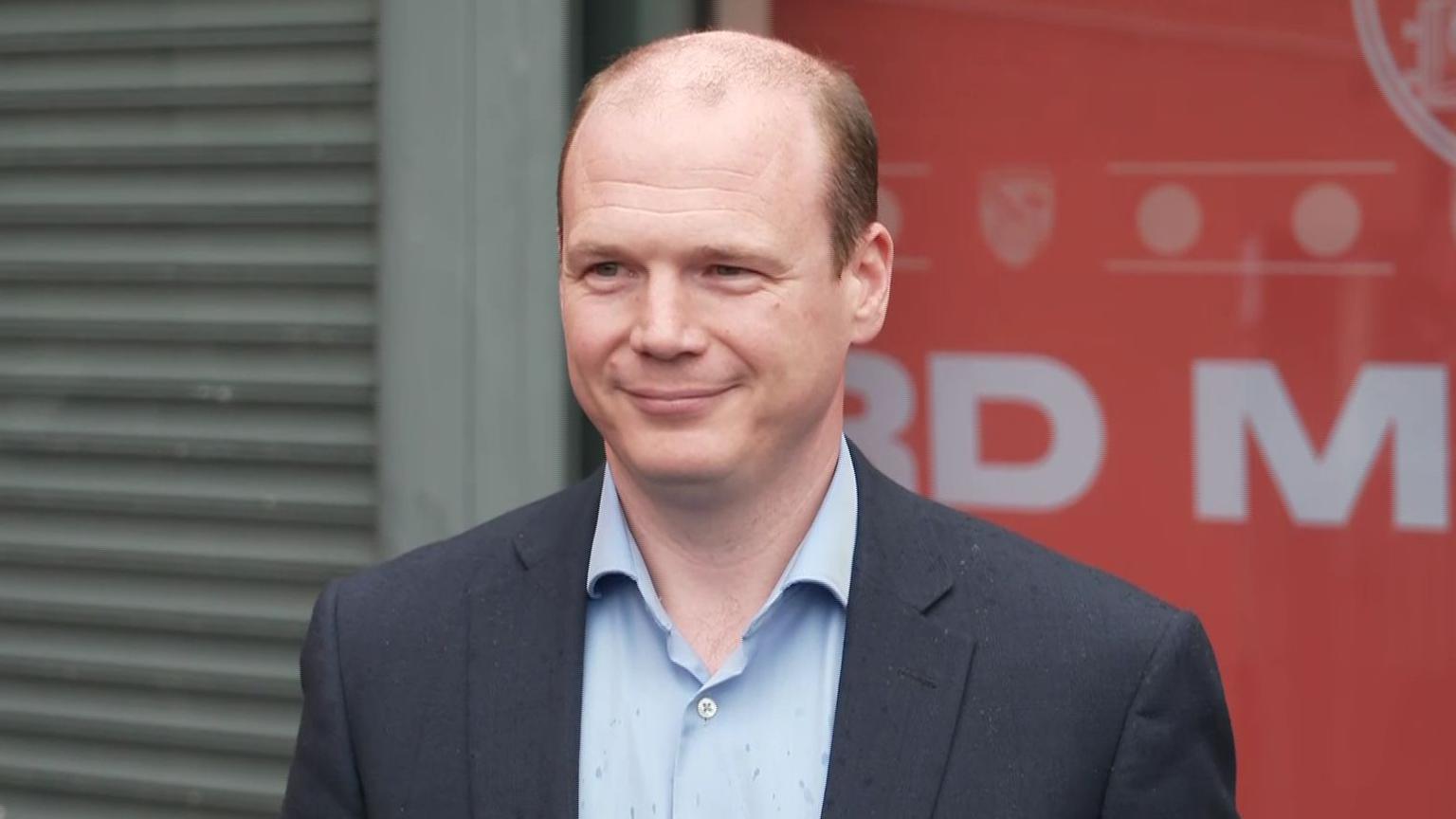 Lyons is wearing a grey blazer and a light blue shirt. He has brown hair around the side of his head and balding on top. He has a slight smile.