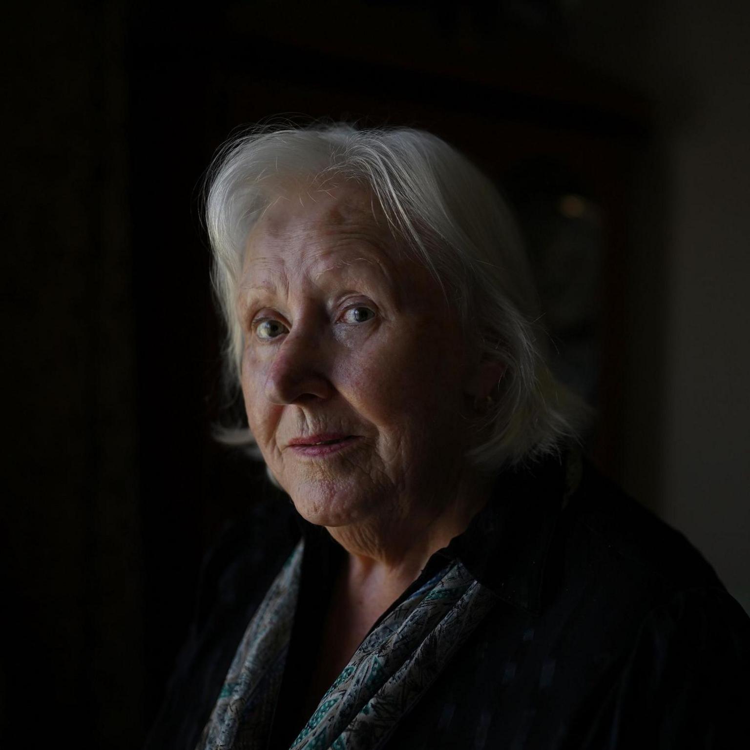 An older woman with medium-length grey hair stands in a living room. We can see her head and shoulders. She is wearing a dark jacket and a patterned silk scarf. In the background, which is out of focus, we can see a picture hanging on the living room wall and a cabinet.