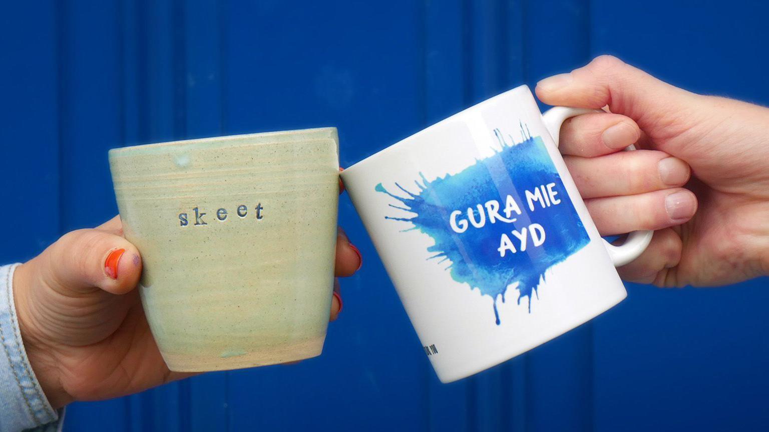 Two hands holding mugs clicking together in front of a blue background. One is green with the word skeet on it, the other is white and blue with gura mie ayd on it.