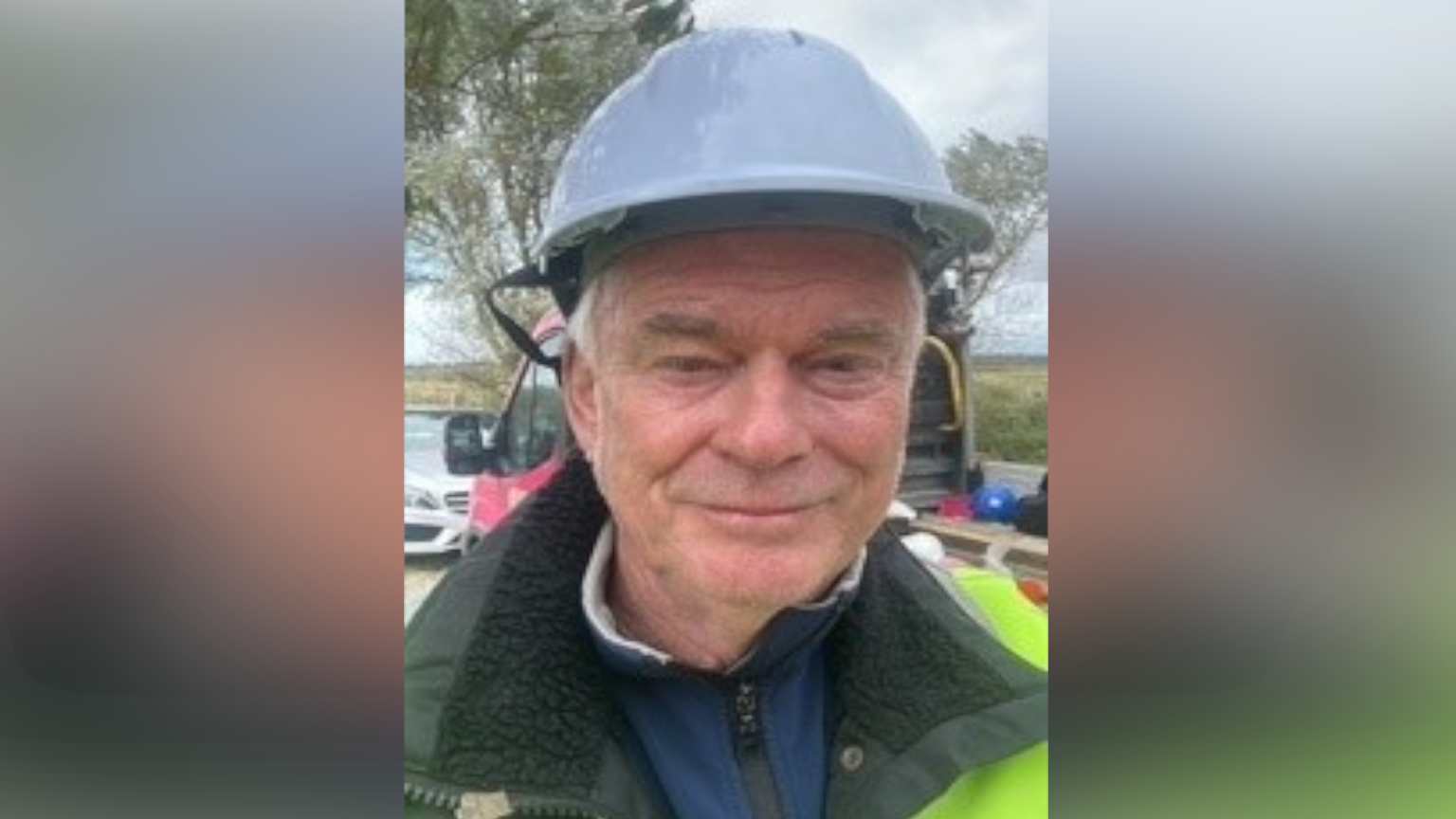 James Forsyth smiles at the camera. He wears a blue hard helmet on his head as well as a hi-vis jacket over a green coat. A flat bed truck can be seen parked behind him.