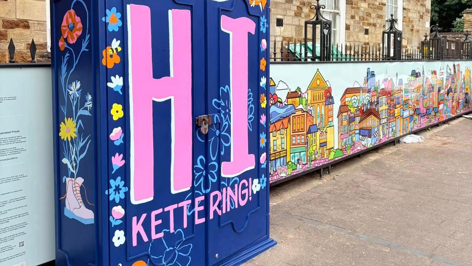 Artwork painted in front of black metal railings, with a box in front, that is painted blue, with colourful patterns and writing on it, that reads "HI Kettering "on it. A brick building is behind the railings.