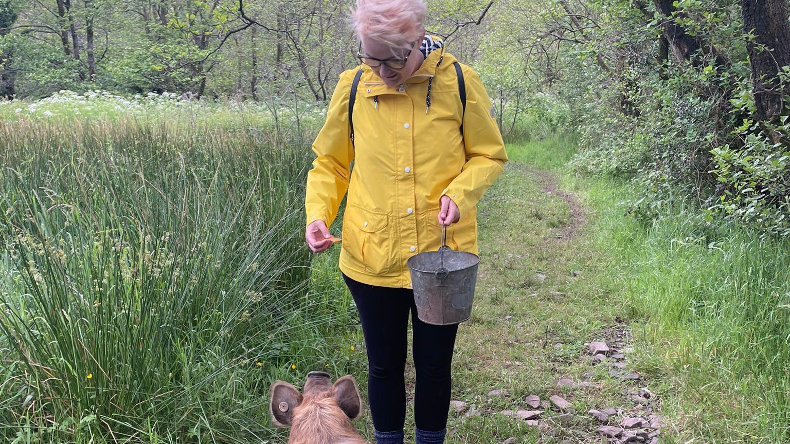 Dr Rebecca Ellis feeding a pig out of a metal bucket, she is in a bright yellow anorak and black leggings and has short pink hair.