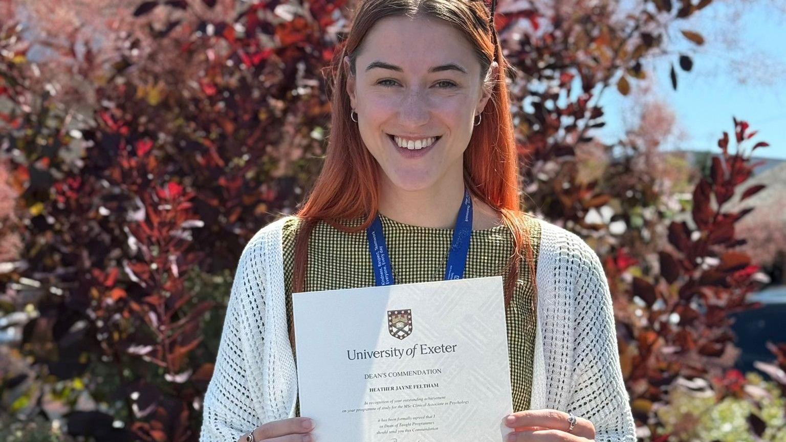 Heather Feltham is standing in front of a red bush holding up a certificate from the University of Exeter. Heather has long, red hair and is smiling.