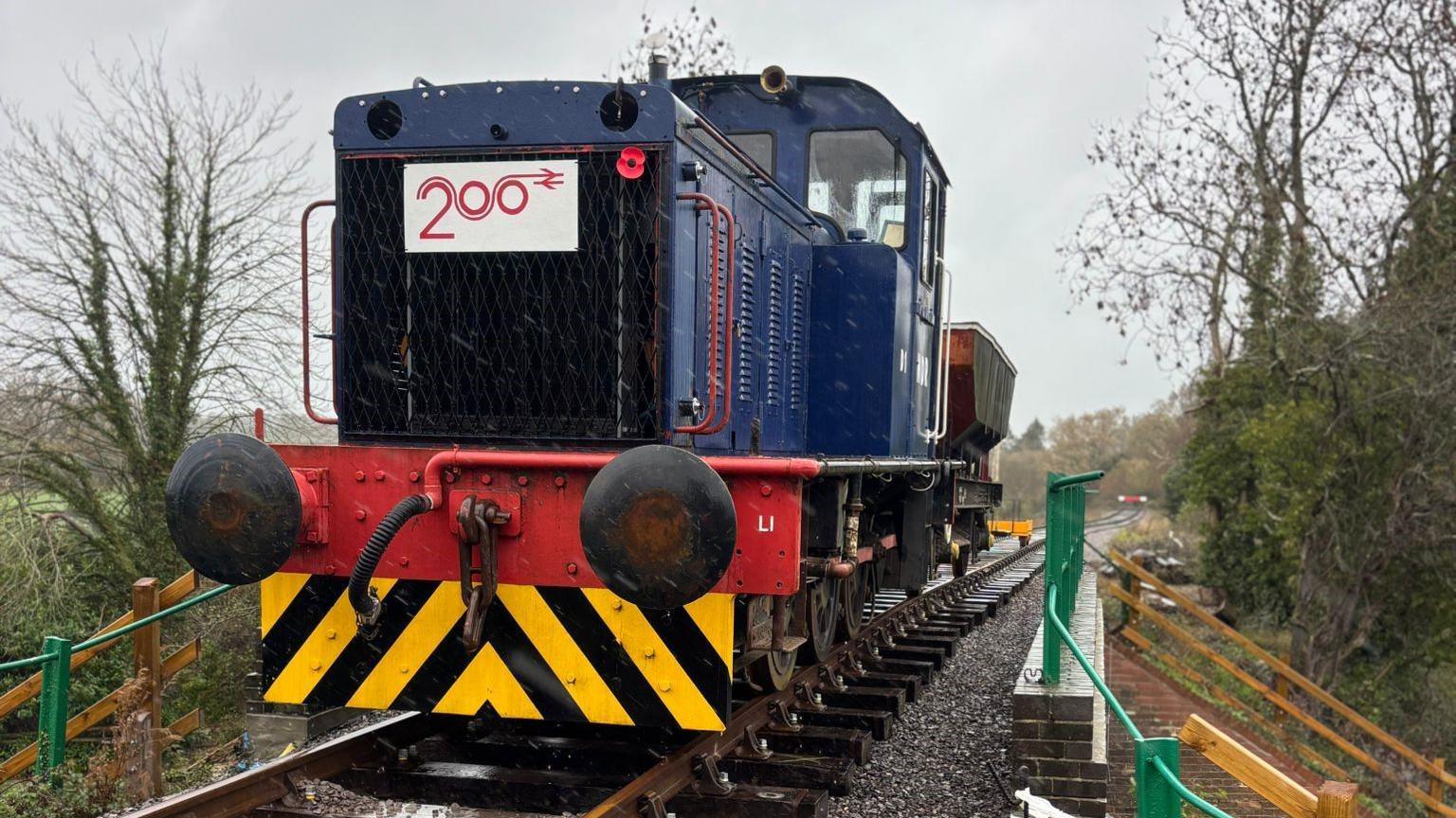 Front view of the blue locomotive standing on the tracks on the bridge. The photographer is standing alongside the track. The front of the loco has a yellow and black striped plate beneath the red buffers. The front of the engine has a Railway 200 logo and a small red Remembrance poppy attached to it. The railings on the bridge are painted green and the track, which is lined by trees, disappears into the distance.