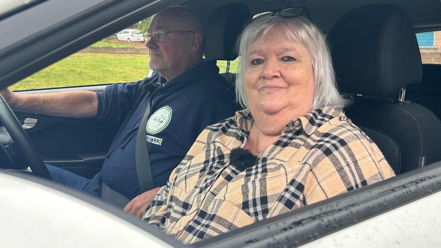 Evie McLean sitting in the passenger seat with volunteer Danny Prew with his hands on the steering wheel. Evie, wearing a checked beige shirt is smiling. 