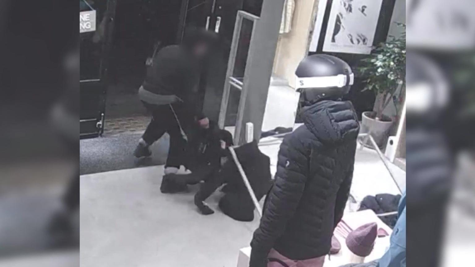 A CCTV image shows a male shoplifter, his face blurred and wearing dark clothing, dragging over a clothing rack, as he pulls items from it