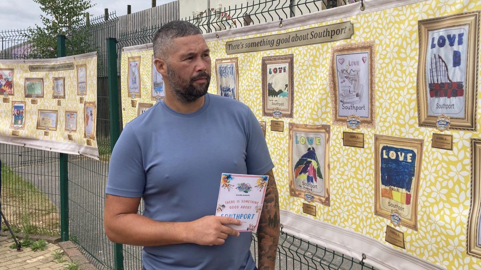 Boxer Tony Bellew poses in front of some of the artwork on display. He wears a light blue t-shirt. The wall behind reads 'There's something good about Southport', underneath some printed pictures of the artwork.