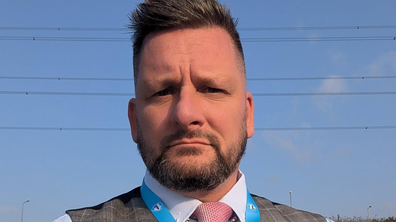 Joel Tetlow has short brown hair and a beard. He is wearing a shirt, waistcoat and pink tie, with the top of a Reform UK lanyard just visible.