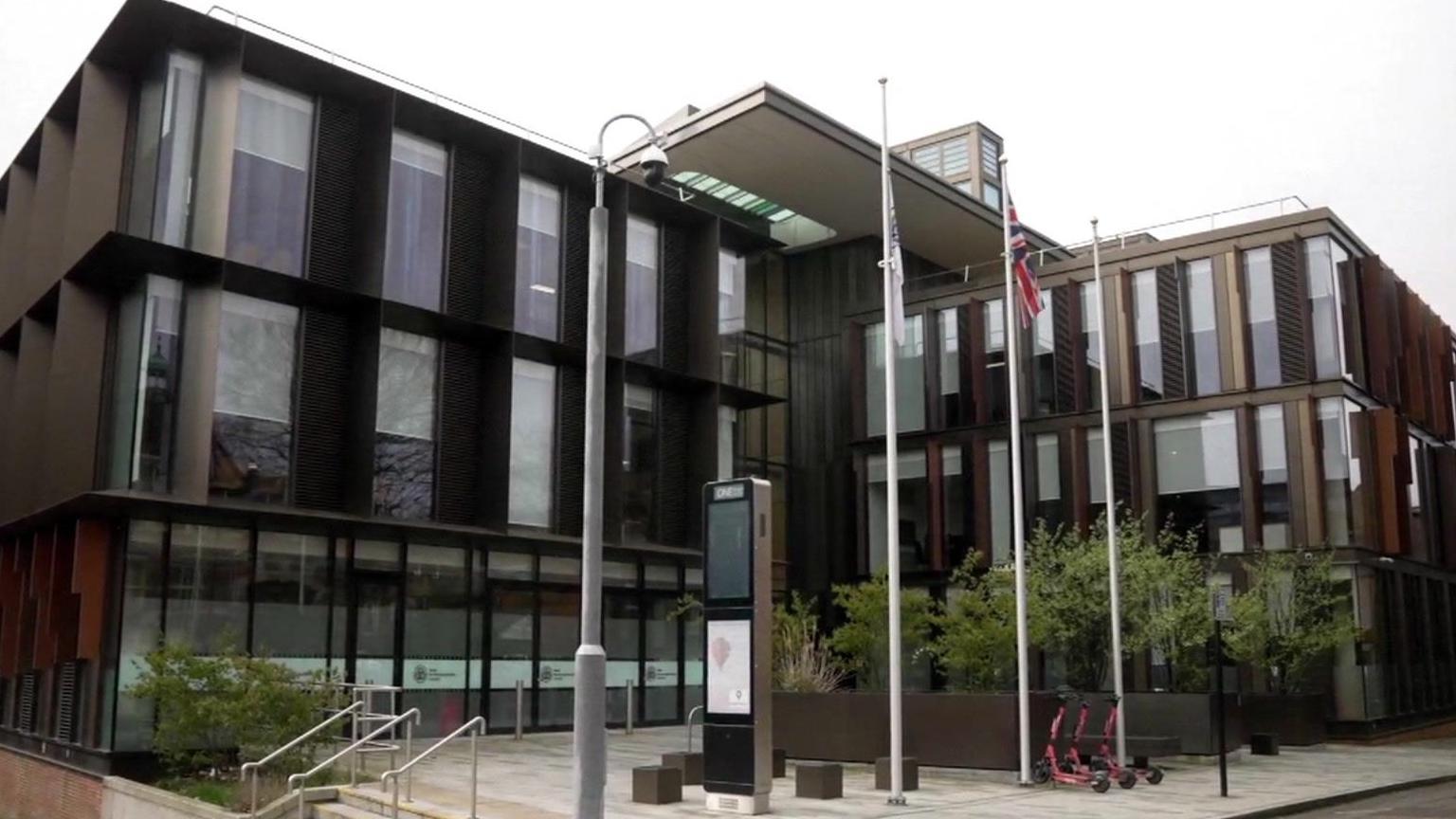 An office building with glass doors, and two rows of vertical windows above the doors. Three flag poles stand in the square at the front of the building. One flies a council flag, the middle flies a Union Jack, and the third is empty. Three red scooters stand on the pavement next to the flag poles. 