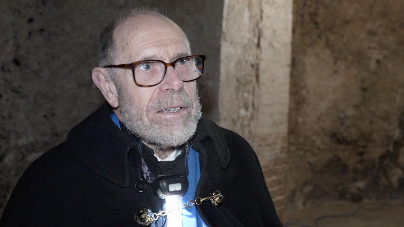 Tony Mills, wearing a black cloak and brown glasses. He is looking off to the side of the camera.