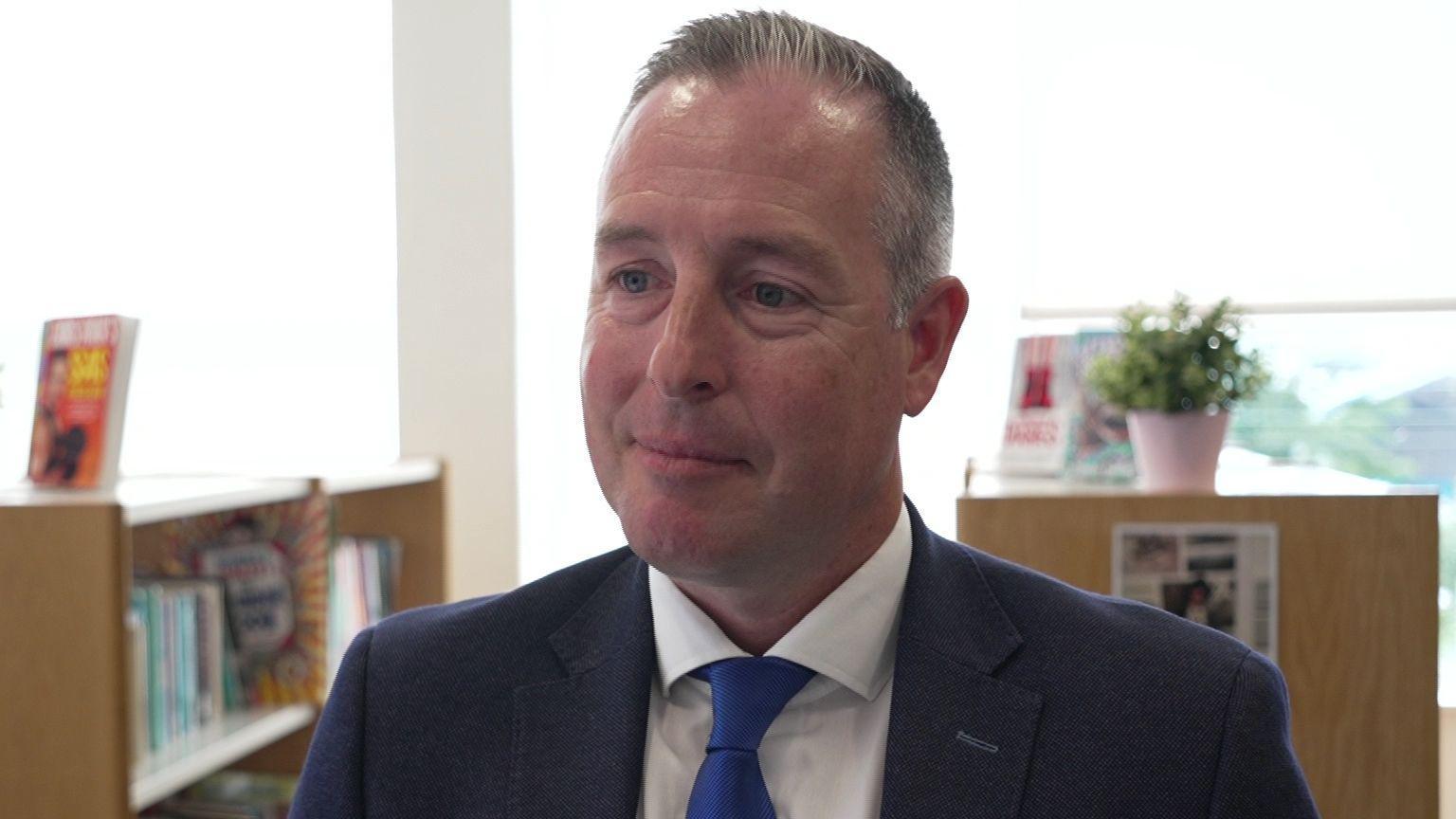 A short grey haired man wears a navy suit with a white shirt and blue tie. Behind him are wooden book shelves. with an array of multicolour titles. There are windows with white blinds behind him, half shut.