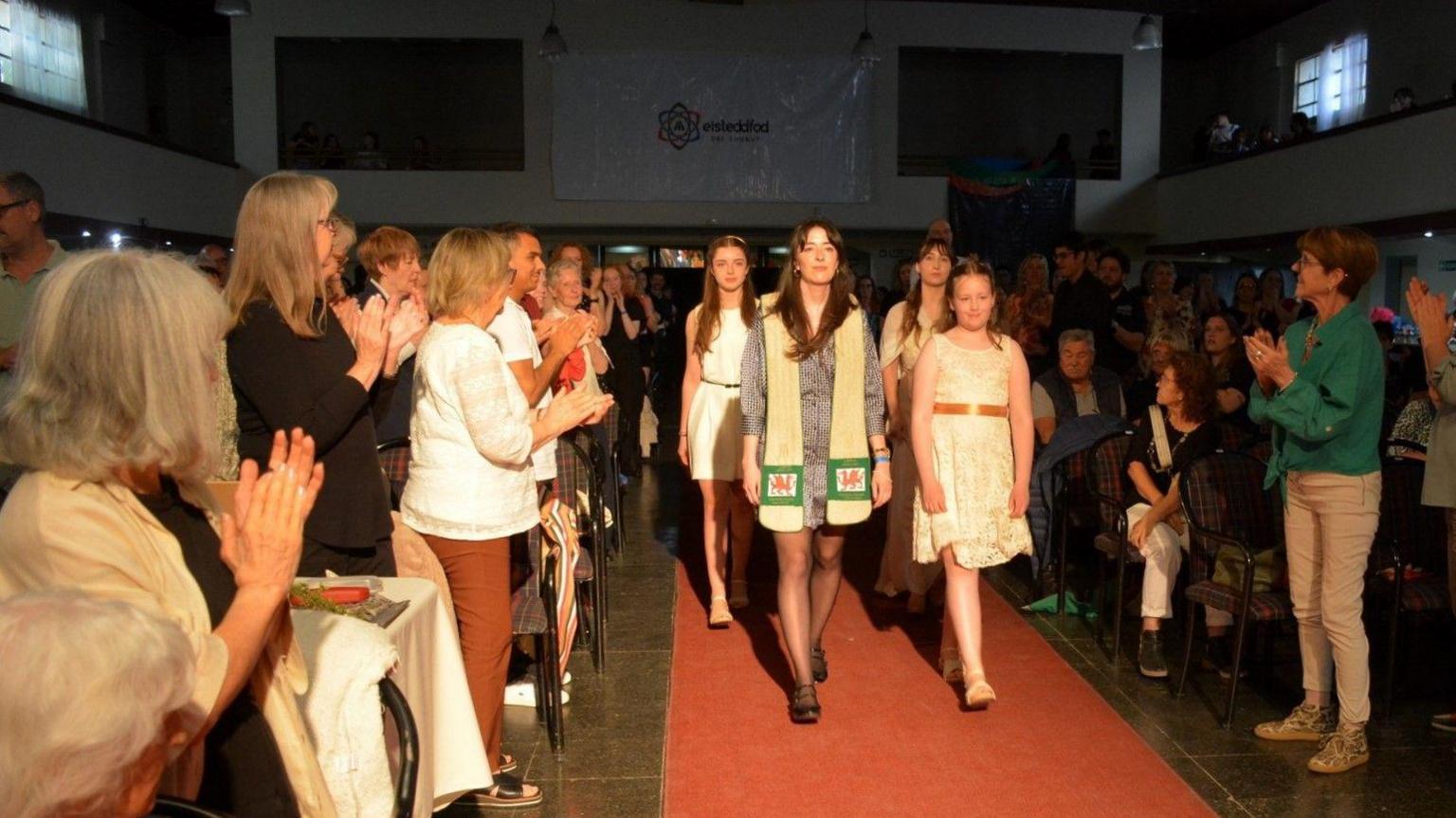 Dynes mewn sgarff efo dreigiau yn cerdded ar garped coch, rhwng gynulleidfa sy'n clapio a gwylio hi.