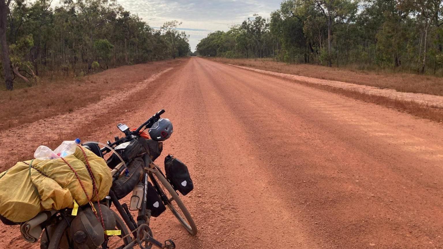 A pushbike packed with luggage leans on its kickstand. It is standing on a wide red gravelly road lined with trees, which stretches into the distance. Apart from the bike, it is empty.