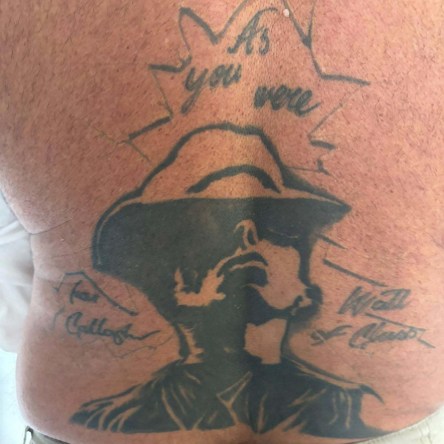 A black and white tattoo of Liam Gallagher which has his name and 'Wall of Glass' written either side of it and As you Were above. It is on John Smallbones' back.