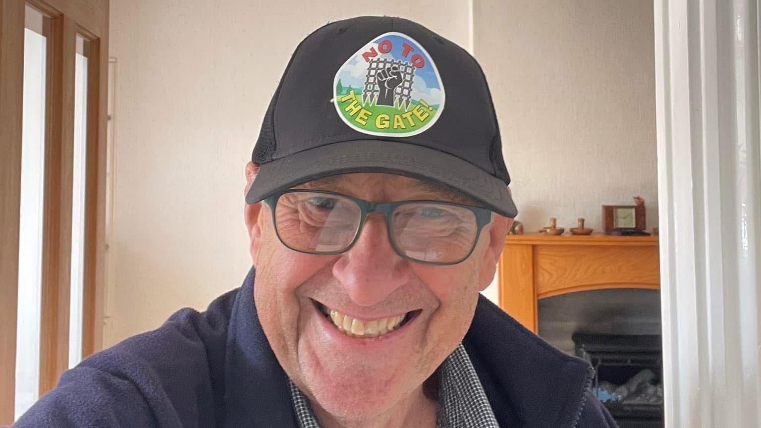 Selfie image of John Warren, aged 73, who wears black rimmed glasses and a baseball cap with a "NO to the gate" sticker on the front. He smiles at the camera. 