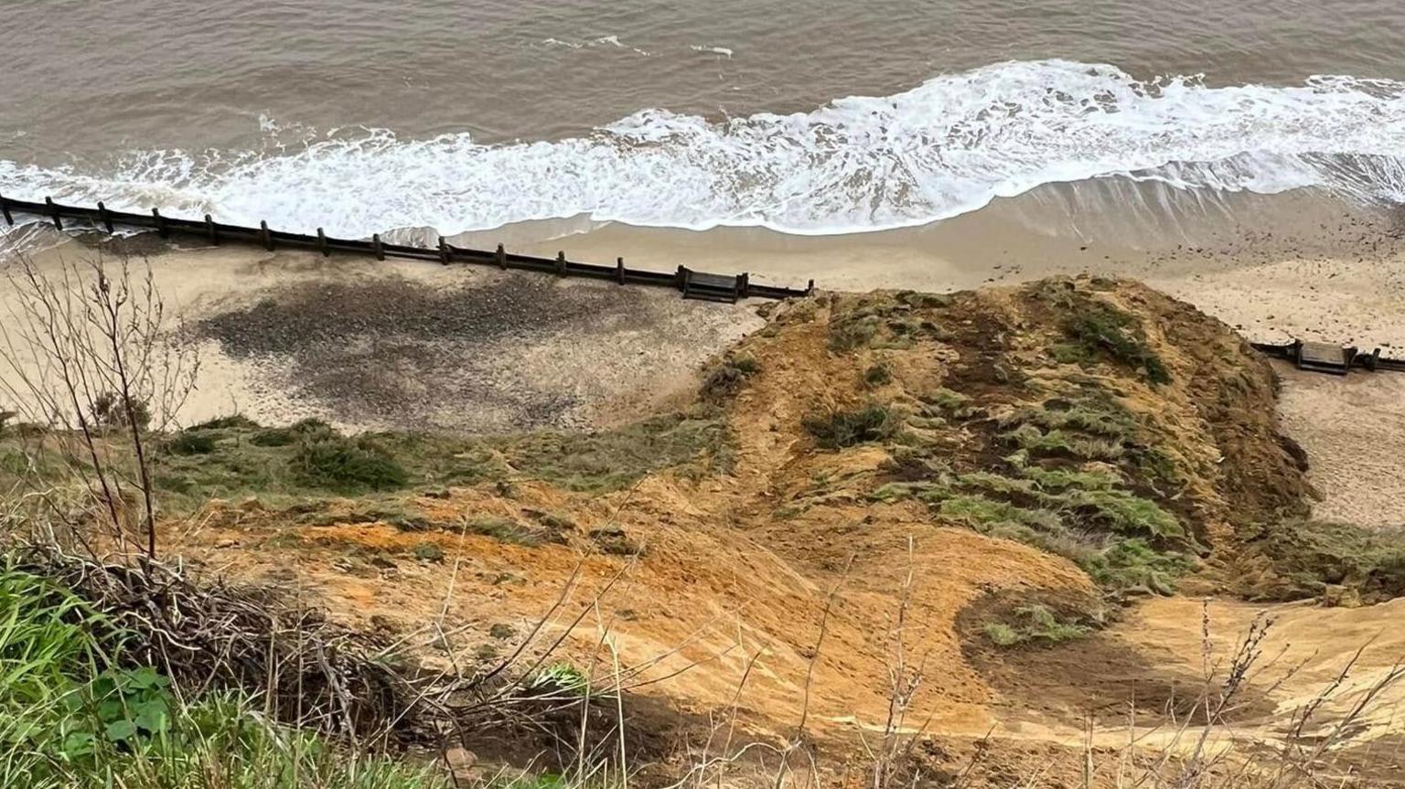 A clifftop view shows where the cliff has fallen on to the beach below. The waves are lapping at the sand