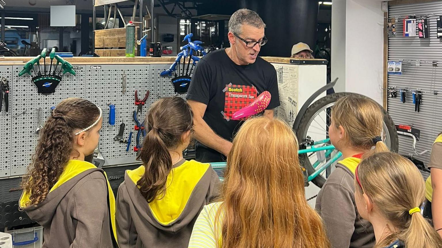 A man and a group of Brownies stand in a bike shop looking at a bike. The man is facing the girls, none of whose faces can be seen, whilst demonstrating something on the bike. The brownies look on in his direction. Behind the man is a wall with a number of tools and a can of spray paint.