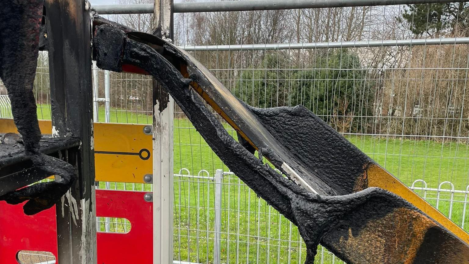 A badly burned yellow slide, the metal of which has become warped and corroded. A fence has been put up around the slide.