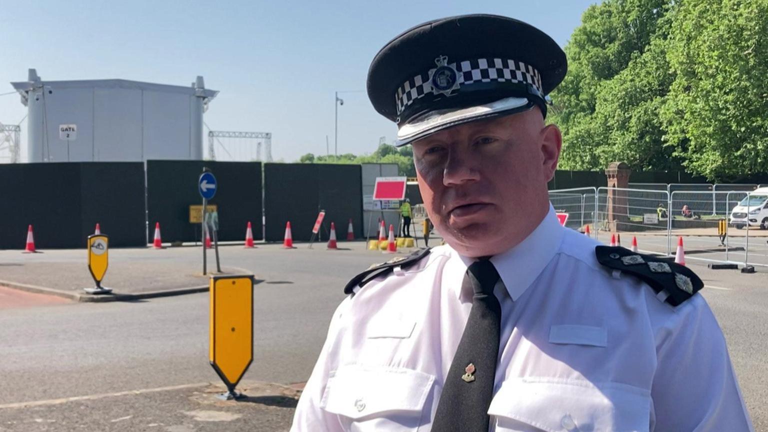 Ch Insp Iain Wyke is wearing a white police uniform shirt with a black tie and a police hat. He is stood at the side of a road at Sefton Park opposite fencing and the back of a large stage. A number of red and white cones and fencing green and metal fencing is in the background.