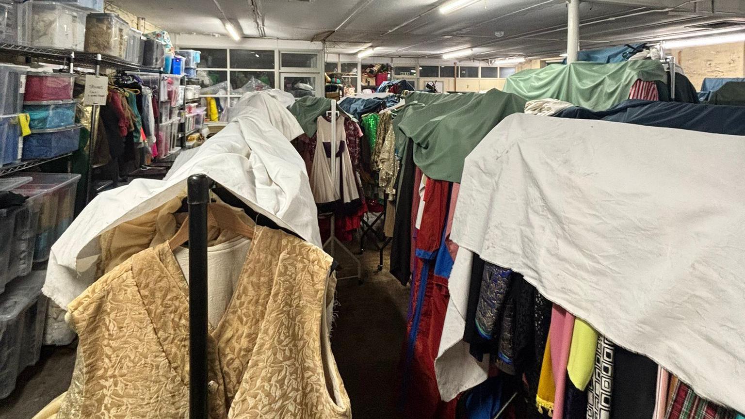 Rows of clothes hangers with colourful clothing costumes, protected by sheets in a large room. Storage boxes to the left and the room is lit by strip lights on the ceiling.