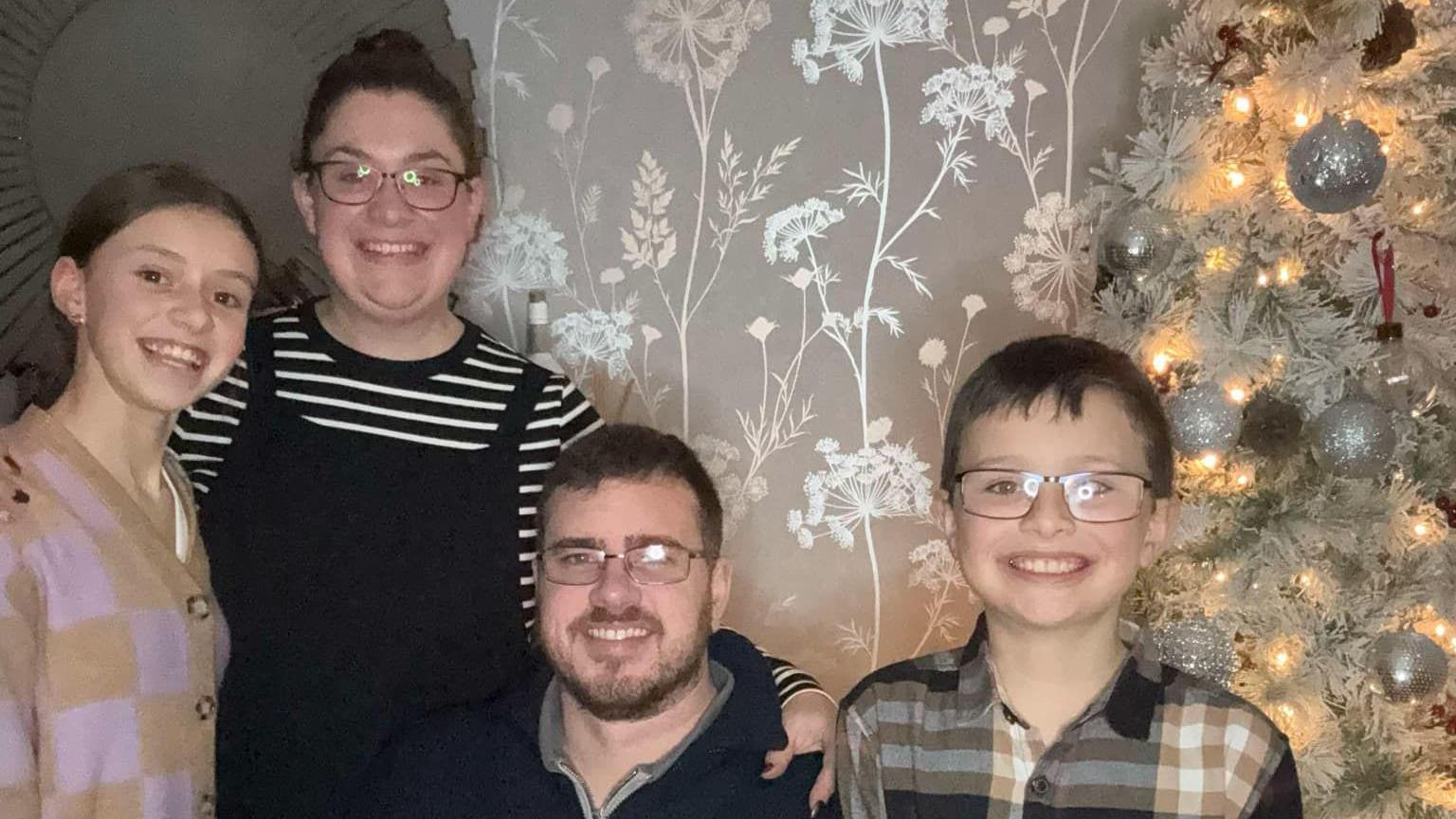 A family photo featuring a mum and dad and their two children. They are standing in a living room next to a tree.