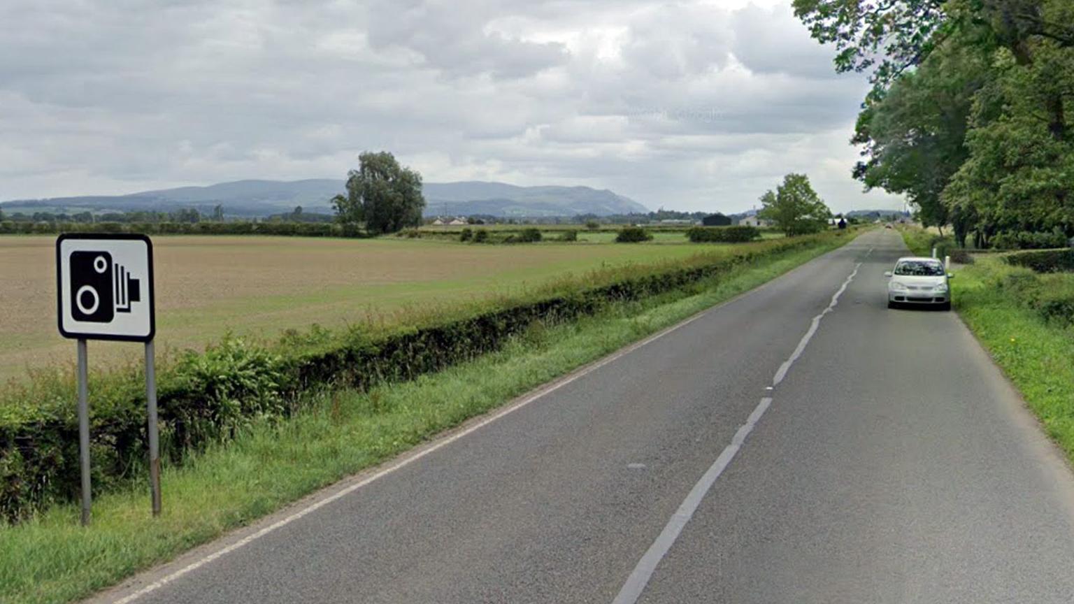 A country road extends into the distance with a silver car and trees and hedges on each side and a speed camera sign to the left.