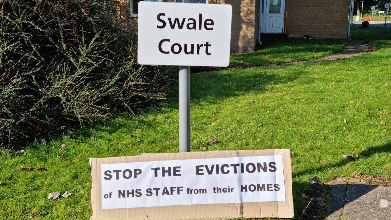 A white post in the grass outside a red brick building reads 'Swale Court'. On the ground is a handmade cardboard sign which reads: "Stop the evictions of NHS staff from their homes."