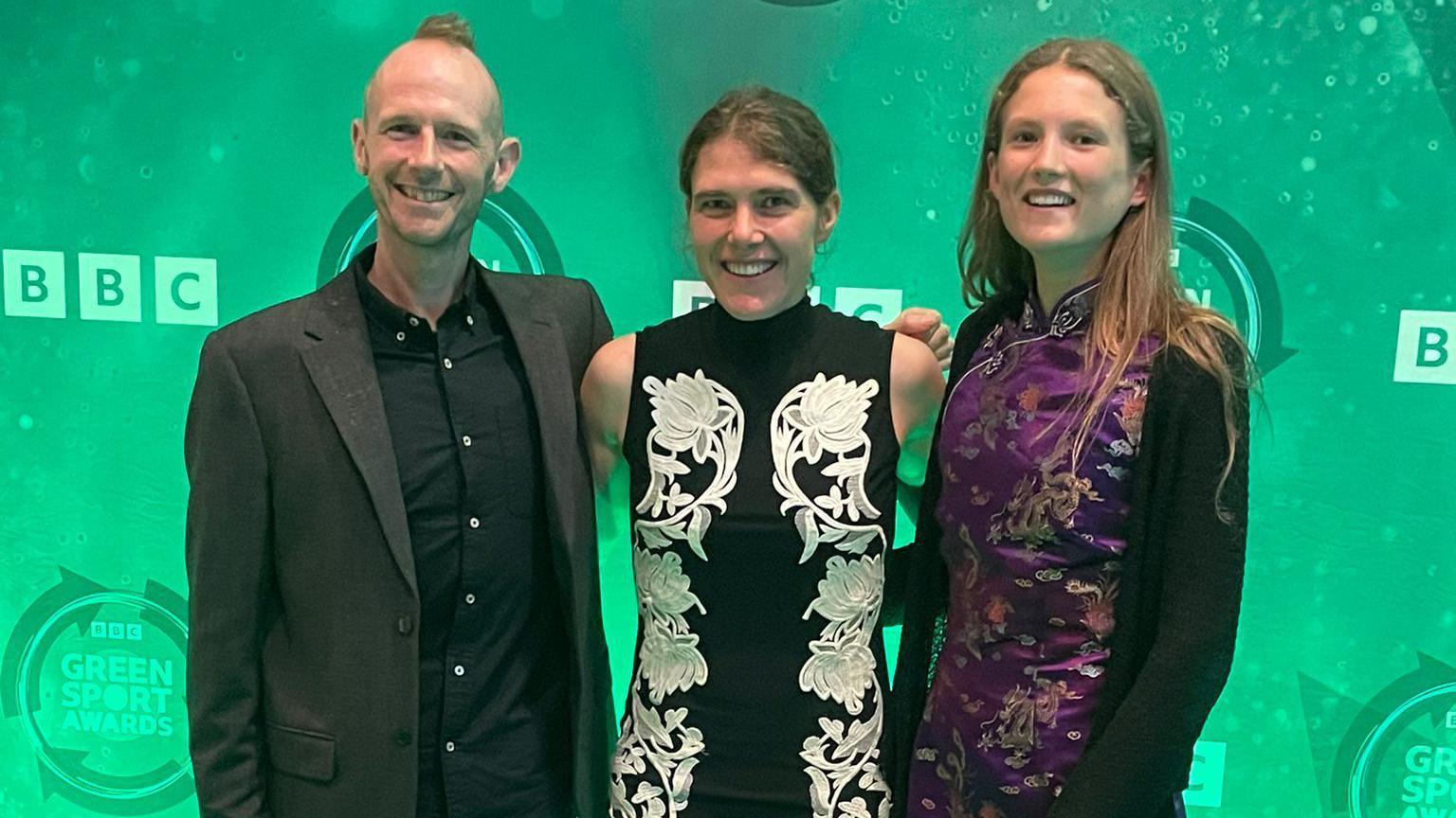 Damian Hall standing with two women at an awards ceremony. They have their arms around each other and are all looking and smiling at the camera